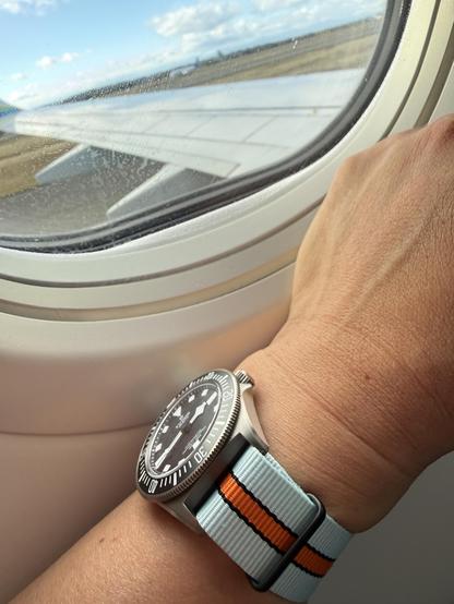A watch on a light blue, orange and black strap held up to an airplane window