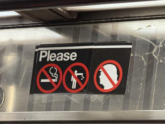 This is a close-up of a sign inside an NYC subway car. It is a standard sign asking people not to smoke or litter. The word “Please” is followed by two icons, both in white silhouette, overlaid by a red circle with a line through it: a smoking cigarette, and a person littering. Some dedicated subversive has pasted on their own custom sticker, thus making a third icon in same style. This third icon is a white silhouette of Trump’s head, with the same red “no” circle over it.