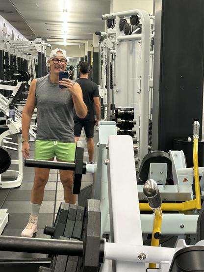 A person in a gym is taking a selfie in front of a mirror. They are wearing a gray sleeveless shirt and green shorts, with a white cap and light-colored shoes. In the background, another individual can be seen, and various gym equipment