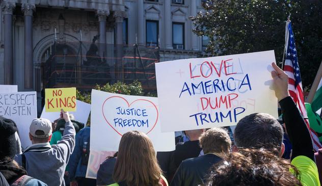 A group of protesters seen from the back.  Visible above them are signs reading "RESPECT EXISTEN", "NO KINGS", "JUSTICE FREEDOM" inside a heart, "LOVE AMERICA DUMP TRUMP" and a US flag on a pole.