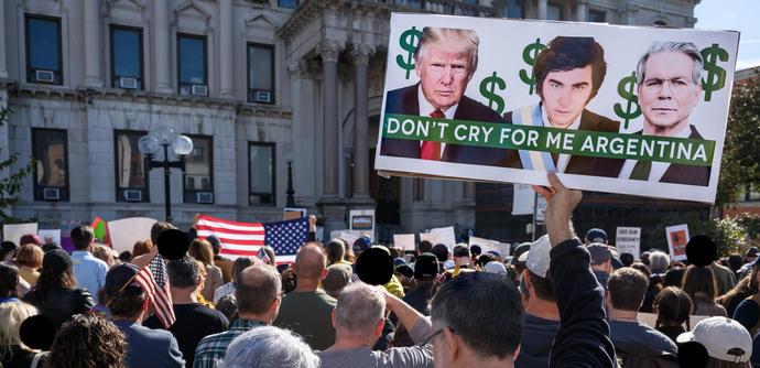 Many protesters seen from behind. We see them holding up US flags and signs.  The largest, most visible sign - nearly filling the upper-right quarter of the frame - shows images of Donald Trump, Javier Milei, and Scott Bessent. There are dollar signs behind them and text running over their chests reading "DON'T CRY FOR ME ARGENTINA".  Some protesters' faces are obscured with black discs.