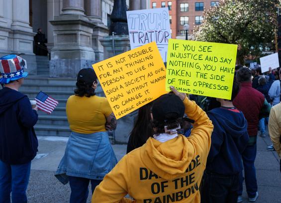 Protesters seen from behind. One, off to the side, wears a bedazzled red white and blue top hat and carries a small US flag.  Closer, and filling much of the frame, someone holds hand-lettered signs on fluorescent card stock (one orange, one yellowish).  One reads "It is not possible for any thinking person to live in such a society as our own without wanting to change it –George Orwell" and the other "If you see injustice and say nothing, you have taken the side of the oppressor. –Desmond Tutu".