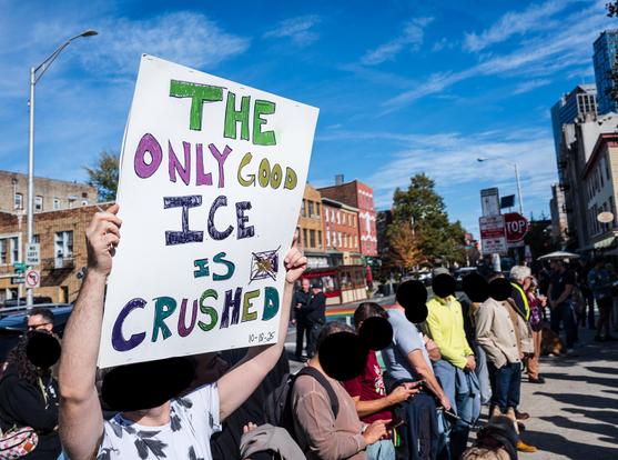 A line of people standing along a sidewalk.  One holds a large sign reading "THE ONLY GOOD ICE IS CRUSHED". Most people's faces are obscured with black discs.