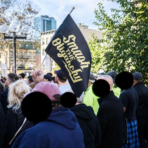 A crown of protesters.  Visible above them, a banner on a staff reads "Smash the Oligarchy" in a stylized script. Protesters' faces are obscured with black discs.