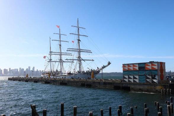 Tall ship docked at the Shipyards Pier. The ship is white and has 3 masts. Downtown Vancouver is in the background.