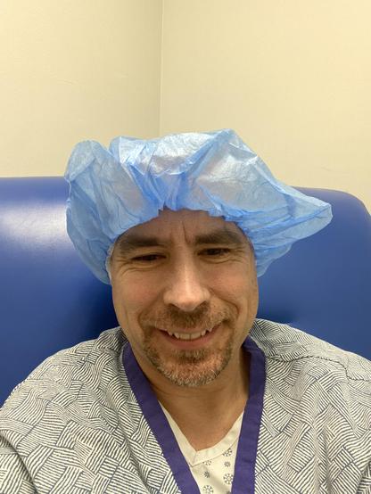 A photo of me smiling nervously wearing a hospital cap, a hospital robe and gown. I’m seated on a blue vinyl high-back chair.