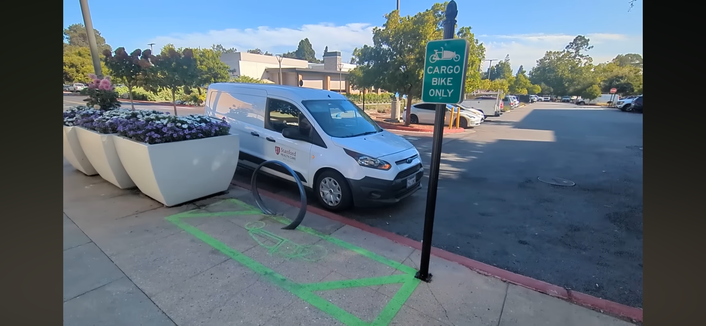 A screenshot of a video showing a “Cargo bike parking” only bike lockup on a sidewalk next to a large car parking lot.