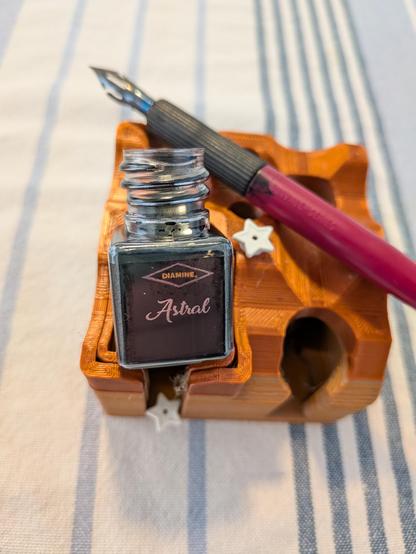 A bottle of Diamine Astral ink sits on a holder, with a couple of Lego stars and a dip pen with a purple handle.