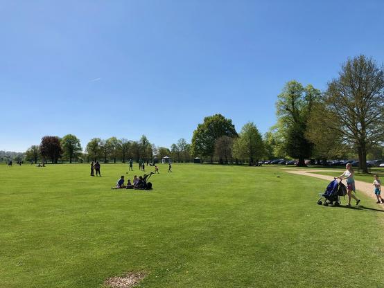 A wide, open grassy field at Belton House in Grantham stretches under a clear blue sky, bordered by rows of leafy trees with spring foliage. Families and small groups are scattered across the lawn, some sitting on the grass while others walk along the dirt path that curves past the tree line. A woman pushes a stroller with a child walking beside her, and more people can be seen in the distance, enjoying the sunny weather. Parked cars are partially visible under the trees to the right, and a small pavilion or shelter is positioned further back along the open space.