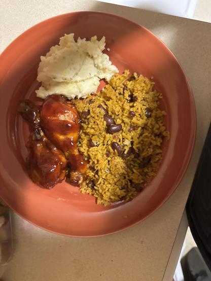A plate of food on a reddish-orange plate. There is a scoop of mashed potatoes in the upper left section, some yellow rice mixed with black or red beans on the right, and two pieces of barbecue chicken drumsticks on the left side of the plate. The mashed potatoes appear creamy, the rice is seasoned, and the chicken is coated in a shiny red sauce. The plate is sitting on a light-colored countertop.

Image description provided with Be My Eyes.