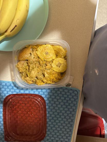 Plastic food container with a meal inside, placed on a counter. The meal includes yellow rice, chicken pieces, and several round fried plantain slices on top, all in a yellow sauce. To the left, there is a light green plate with bananas, and below the container is a woven blue mat with a closed red container on it. Part of someone's clothes and a bit of a red object are visible on the right side of the image.

Image description provided with Be My Eyes.