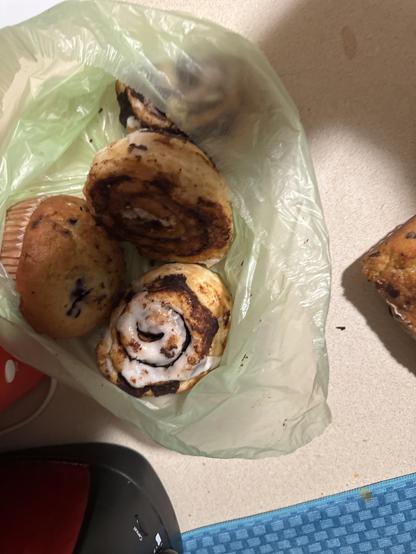 Baked goods in a light green plastic bag. The bag contains two cinnamon rolls with icing and what appears to be two blueberry muffins. The items are on a light countertop, with a blue textured mat and a piece of another baked good visible on the side. There is also part of a red and black kitchen appliance in the lower part of the image.

Image description provided with Be My Eyes.