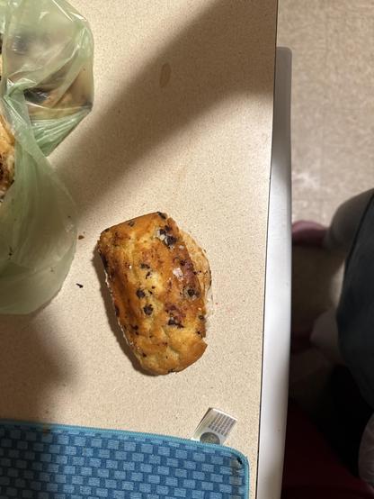 Baked pastry with chocolate chips, resting on a light-colored countertop. The pastry looks rectangular and golden-brown, with small dark chocolate chips scattered on top. There is a green plastic bag with more baked goods partially visible to the upper left and the corner of a blue textured cloth or mat at the bottom. Near the blue mat, there's a small white and green tear-off packet. At the right side of the image are two legs, one wearing a pink slipper and the other in a gray garment.

Image description provided with Be My Eyes.