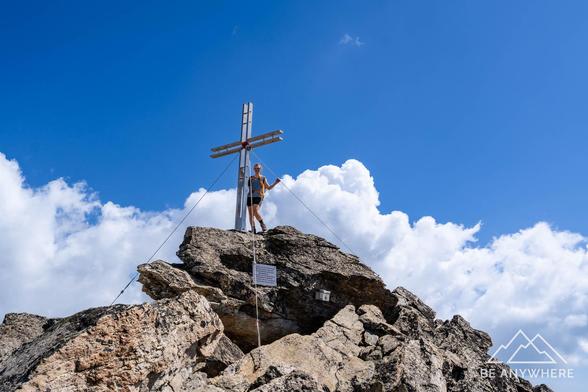 A hiker standing by the large summit cross of the Almagellerhorn under a blue sky with scattered clouds, a moment of quiet triumph on the rocky peak.