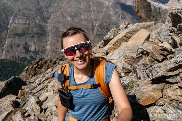 Close-up of a smiling hiker wearing sunglasses and an orange backpack against a rugged alpine backdrop.