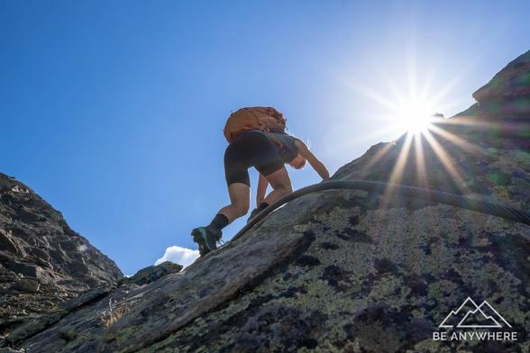 Low-angle shot of a hiker climbing up sunlit rocks — the bright sun flaring behind, capturing the power and beauty of the ascent.