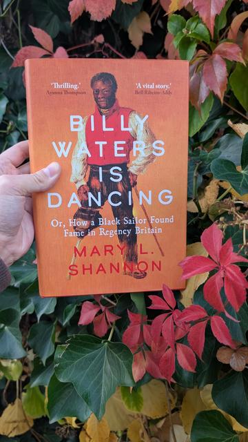 Holding the book "Billy Waters is dancing" in front of a wall overgrown with autumnal leaves and vines