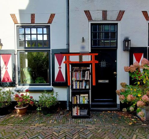 Sfeervol tafereeltje met een oude, wit gepleisterde woning met een donkergroene houten deur, links en rechts daarvan een raam met groene kozijnen en meerdere kleinere ruitjes, met opzij opengeslagen rood-wit houten luiken. Voor het huis verschillende planten in terracota en zwarte potten.
Daarvoor staat een hoge langwerpige rood wit zwart houten kast met glazen deur met vijf etages boeken erin. Aan de bovenkant in het midden een zwart houten langwerpig bordje met Japanse
tekens in geel. (Zie vlg alttekst)
Links schijnt de zon op de muur. De rest is in de schaduw. 

Dit alles op een oud sfeervol klinkerstraatje.