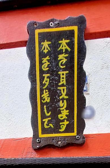 Ingez👀md op het bordje: zwarte schulprand aan buitenzijde, daarbinnen een geel smal kader er dáárbinnen de Japanse tekens in geel, in twee verticale kolommen.  De vertaling is ongeveer (lezen: eerst de rechterkolom van boven nr beneden en dan idem de linkerkolom): 

Ik 
neem 
boeken 
in

en 
deel
boeken 
uit