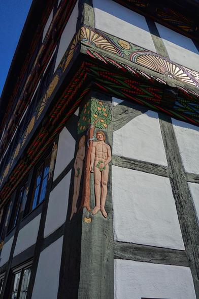 Close-up of the Adam and Eve House in Höxter, Germany, showcasing its distinctive half-timbered architecture. The building features a richly decorated wooden beam with vibrant, intricate patterns and carvings. A relief sculpture of Adam and Eve stands beneath a tree with fruit, symbolising the biblical scene. The white plaster walls and dark wooden beams are characteristic of traditional German timber-framed buildings, set against a clear blue sky.