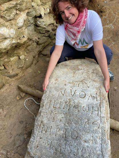 Una donna sorridente posa accanto a una stele funeraria romana con iscrizioni visibili, situata in un'area di scavo archeologico.