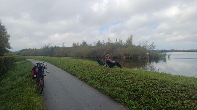 twee ligfietsen staan geparkeerd langs een fietspad. rechts van het fietspad een dijkje. op het dijkje zit een dame op een campingstoeltje, uitkijkend op het water. het tweede stoeltje is leeg.