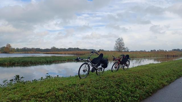 Een rode Chamsin en witte Gaucho ligfiets poseren op een dijkje bij de Nieuwkoopse plassen. op de achtergrond water, nog een dijkje, nog meer water en een kalende boom aan de horizon. grijs-blauwe lucht die ook weerspiegelt in het water.