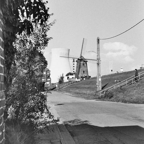 Black & White photo showing a windmill in front and the gigantic cooling towers of a nuclear power plant at the back. Left side of the image is dominated by a brick wall and some vegetation close-up. The windmill comes out dark while the gigantic cooling tower is slightly over-exposed and almost fades in to the light sky.