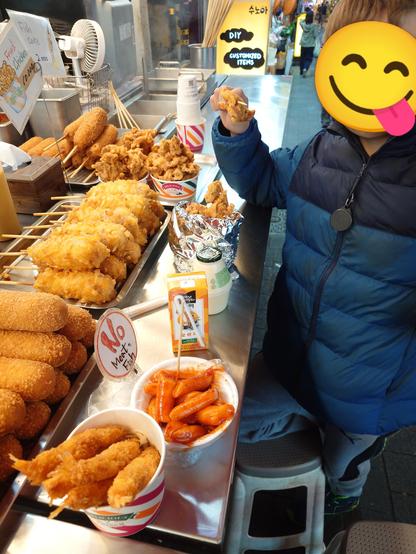 A street vendor counter with visible corn dogs, cheese corn dogs and fried chicken.
My kid is eating a piece of fried chicken from a big bowl. Next to it some orange juice, my tteokbokki and fried shrimps.