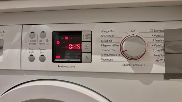 Frontansicht einer Waschmaschine. Das Bedienfeld zeigt ein digitales Display mit rot leuchtender Restzeit „0:38“ und mehreren Symbolen für Waschprogramme. Rechts befindet sich ein Drehregler für verschiedene Programme wie „AllergiePlus“, „Sportswear“, „Extra Fein“ und „Sanft Schleudern“. Einige Funktionstasten links des Displays sind beschriftet mit „Bügel-leicht“, „Spülen plus“, „Speed Perfect“ und „Eco Perfect“. Auf der rechten Seite ist ein Stück Klebeband zu sehen.