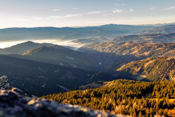 Das Bild zeigt eine atemberaubende Berglandschaft im warmen Licht der goldenen Stunde spät am Nachmittag. Die Sonne steht tief und wirft lange Schatten, wodurch die Konturen der Hügel und Täler besonders hervorgehoben werden.
Zu sehen sind mehrere Ebenen bewaldeter Hügel. Die Landschaft ist von einer Mischung aus Nadel- und Laubbäumen bedeckt, was für eine abwechslungsreiche Textur sorgt. In der Ferne liegt ein sanfter Schleier aus Nebel der dem Bild Tiefe und eine mystische Atmosphäre verleiht.
Der Himmel ist überwiegend klar mit ein paar zarten, federartigen Wolken. Das warme Sonnenlicht betont die Höhen und Tiefen der Landschaft und verleiht dem Bild eine ruhige, fast meditative Stimmung.
