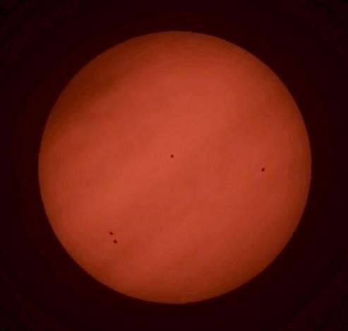 Obraz przedstawia duże, czerwone ciało niebieskie, a raczej pomarańczowe Słońce, z kilkoma mniejszymi ciemnymi plamami widocznymi na jego powierzchni.