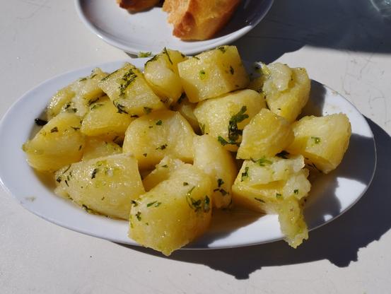 Un plato blanco lleno de patatas cocidas y cortadas en trozos grandes, de color amarillo claro. Las patatas están cubiertas con aceite de oliva brillante y espolvoreadas con perejil fresco picado, que se ve como pequeños puntos verdes. Se encuentran sobre una mesa blanca. En el fondo, parcialmente visible, hay otro plato blanco con trozos de pan. La escena está iluminada por una luz brillante que proyecta sombras nítidas sobre la mesa.