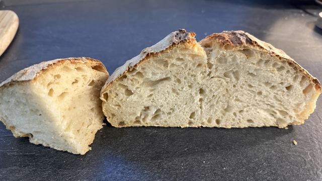 Brotlaib, aufgeschnitten, mit luftiger, poröser Krume und knuspriger Kruste, liegt auf einer dunklen Oberfläche.