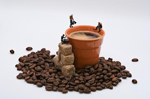 A coffee cup filled with dark coffee sits atop a mound of coffee beans, surrounded by sugar cubes. Three miniature divers figures are positioned on the cup and nearby.