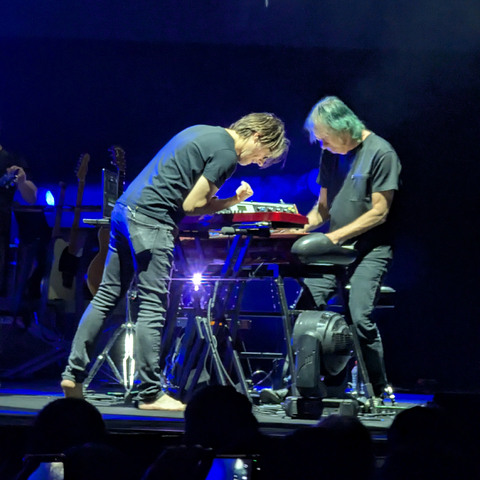 Una imagen de dos músicos actuando en un escenario oscuro con iluminación azul profunda. A la izquierda, un hombre joven de pelo castaño claro, vestido con camiseta y pantalones vaqueros negros, está descalzo y se inclina intensamente sobre un teclado electrónico rojo y negro. Su mano derecha está cerrada en un puño mientras mira el teclado. A su derecha, un hombre mayor con pelo largo, que parece verde o azul debido a la iluminación del escenario, está sentado en un taburete. Él también viste una camiseta y pantalones vaqueros negros y se inclina sobre el teclado, con las manos sobre las teclas. Un foco de luz brillante emana de debajo del soporte del teclado. En el fondo borroso, se aprecian siluetas de guitarras en soportes y, en la parte inferior de la imagen, las cabezas y teléfonos de la audiencia.