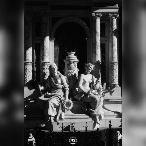 Black and white photo of elaborate grave ornaments. It features two bare-chested female figures, representing the arts and progress.