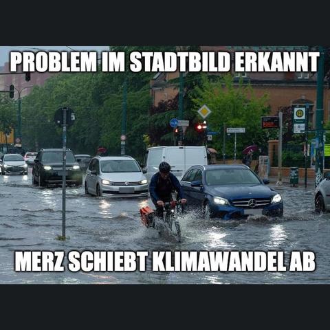 Das Bild zeigt eine Strassenszene mit mehreren Fahrzeugen in einer überfluteten Strasse. Mehrere Autos stehen im Wasser, darunter ein schwarzes Auto, ein silbernes Auto und ein weisser Transporter. Eine Person auf einem Fahrrad fährt durch das Wasser, wobei das Wasser spritzt. Im Hintergrund sind Gebäude und Strassenlaternen zu sehen. Auf dem Bild befindet sich Text: „PROBLEM IN STADTBILD ERKANNT“ und „MERZ SCHIEBT KLIMAWANDEL“.