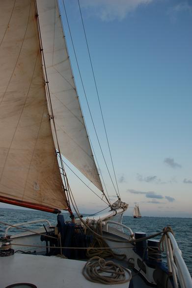 photograph shows part of sails and bow of a sailing boat. There is another boat visible ahead of this one.