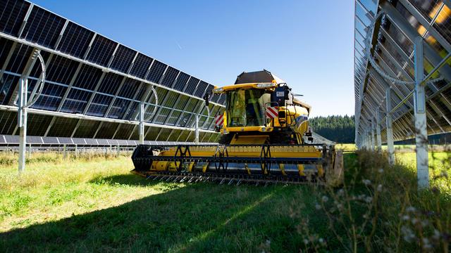 Strahlend blauer Himmel. Darunter ein herbstliches Feld. Im Bild-Mittelpunkt steht ein gelber Mäh-Drescher unter zwei Reihen PhotoVoltaik. Die Solar-Panele sind dem Mäh-Drescher abgeneigt, um möglichst viel Platz für die Agrar-Maschine frei zu geben.