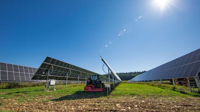 Strahlend blauer Himmel. Darunter ein herbstliches Feld. Zu sehen sind mehrere Reihen PhotoVoltaik-Tracker (= bewegliche, der Sonne nachlaufende Solar-Tische) und zum Größen-Vergleich ein klein wirkender Traktor.