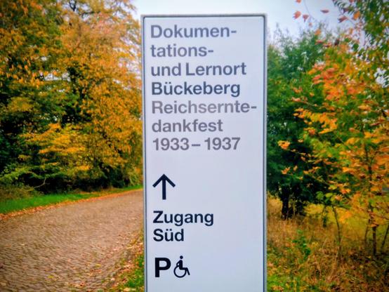 Wegweiser zum Dokumentations- und Lernort Bückeberg am oberen Zugang zum Gelände neben einem Kopfsteinpflaster-Weg; im Hintergrund Herbstlaub.
