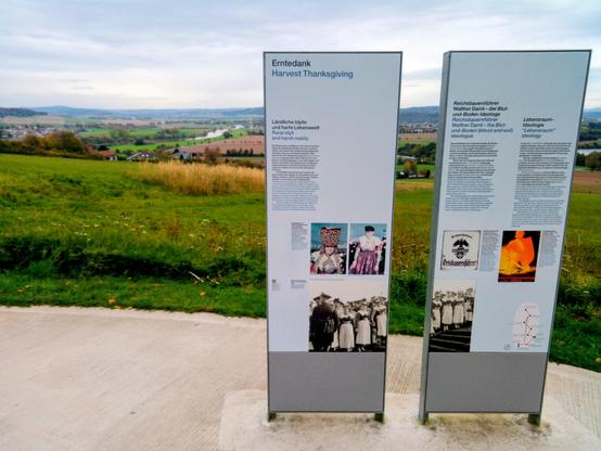 Zwei Ausstellungstafeln mit Texten und Abbildungen zu Themen wie "Erntedank", "Blut-und-Boden-Ideologie" und "Lebensraum-Ideologie" im #Nationalsozialismus; mit Blick über den Hang hinunter in die Landschaft und auf die Weser im Hintergrund.