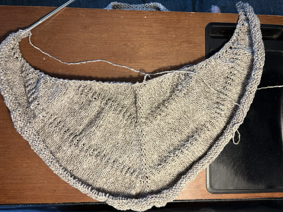 A knitting project lies on a wood-tone lap desk. It is knit in a heathery light-brown and beige wool yarn, and is beginning to show its triangular shawl shape, with three stripes of stockinette stitches separated by thin stripes of twisted stitch ribbing.