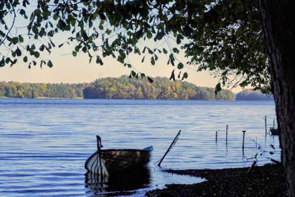 Das Bild zeigt einen See in einem Waldgebiet. Man blickt von der schattigen Seite auf die sonnige, bewaldete Seite gegenüber. Am diesseitigen Ufer ist ein Boot festgemacht.