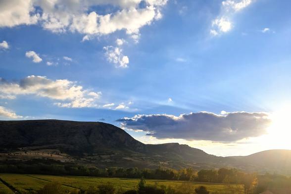 Picture of landscape at sunset with mountain range in background, and valley with row of trees and green field in foreground; sun rays visible on right of frame through partially cloudy blue sky