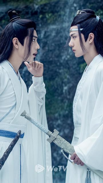 An image focused on teen Wei Wuxian and Lan Wangji standing face to face. Wuxian's left hand is raised with his thumb extended to point behind him, his mouth open as he speaks to Wangji about what he is gesturing toward. Wangji listens with his customary icy expression.