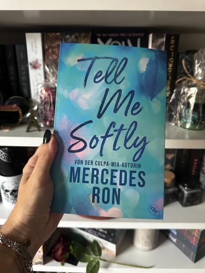 Ich halte das Buch Tell me Softly von Mercedes Ron vor meinem Bücherregal in meiner Hand.

Das Cover des Buchs wird gezeigt.