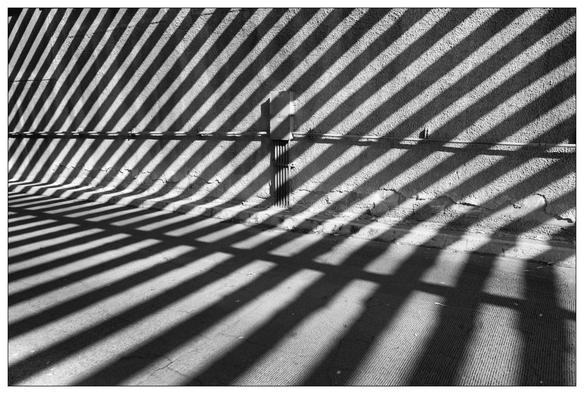 Black and white photo of strong diagonal shadows cast by a fence on a textured ground with a singular utility box and conduit centrally positioned, creating contrast between light and shadow.