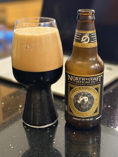 A stout glass half full of dark beer and half full of thick tan head. The short brown bottle has a black and gold label with a stencil of Rasputin on its middle.